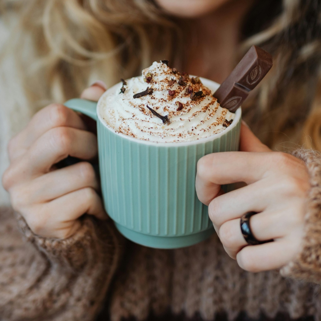 Person holding a green mug with hot chocolate and whipped cream. Mugs of hot chocolate with toppings beside herbal teas and fruit juices.