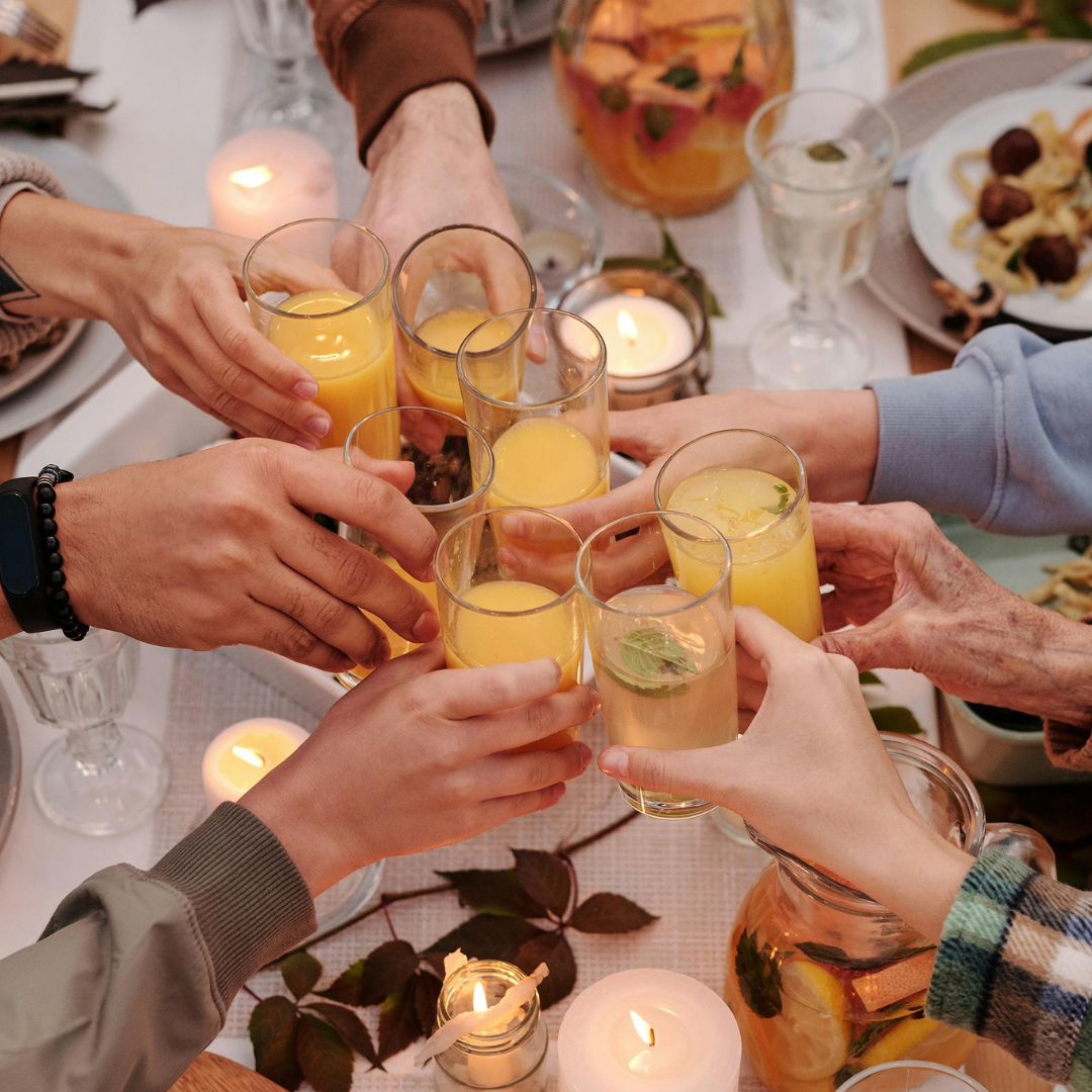 People toasting with glasses of orange juice at a festive table with candles and food. A cozy dinner table with colourful plant-based dishes inspired by world cuisines.
